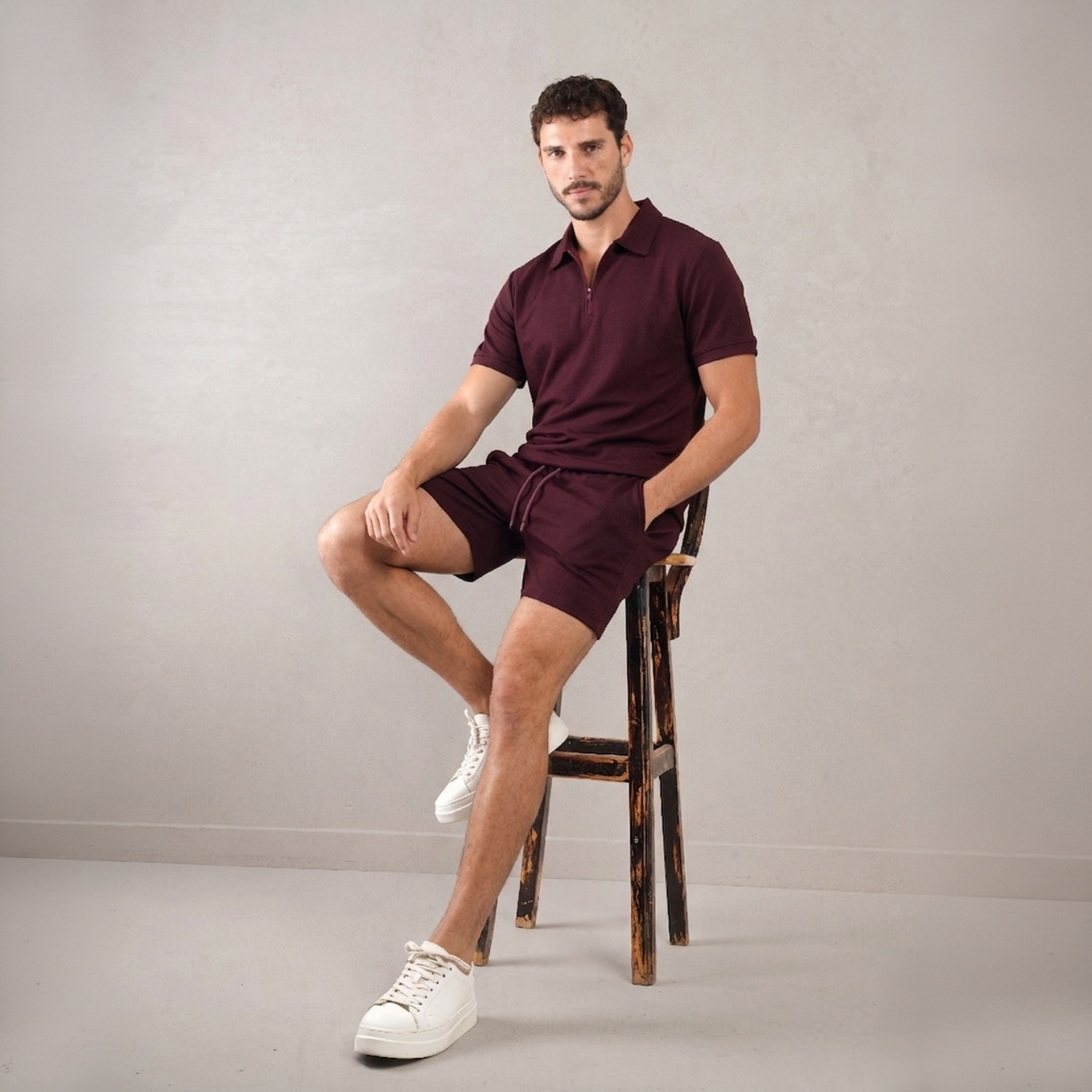 Man sitting on a wooden stool wearing a maroon polo shirt and shorts with white sneakers against a plain background.